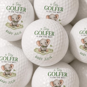Könnte beinhalten: Weiße Golfbälle mit einer aufgedruckten Illustration eines grauen Babyelefanten, der einen Golfschläger auf einem grünen Grasfleck hält. Der Text lautet a tiny GOLFER IS ON THE WAY! und BABY JULIE in dunkelgrüner und schwarzer Schrift.