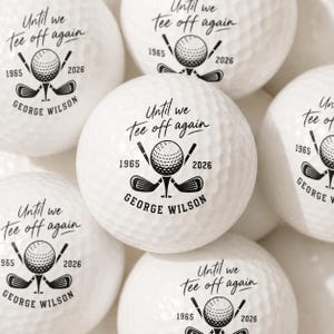 May include: A collection of white golf balls featuring a black printed graphic of a golf ball on a tee crossed with two golf clubs. The text reads "Until we tee off again", "1965", "2026", and the name "GEORGE WILSON" in a circular layout.