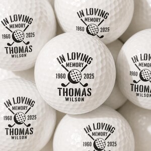 May include: A collection of white golf balls featuring a black printed design. The text reads IN LOVING MEMORY, 1960, 2025, and THOMAS WILSON, accompanied by a graphic of two crossed golf clubs and a small golf ball in the centre of the text.