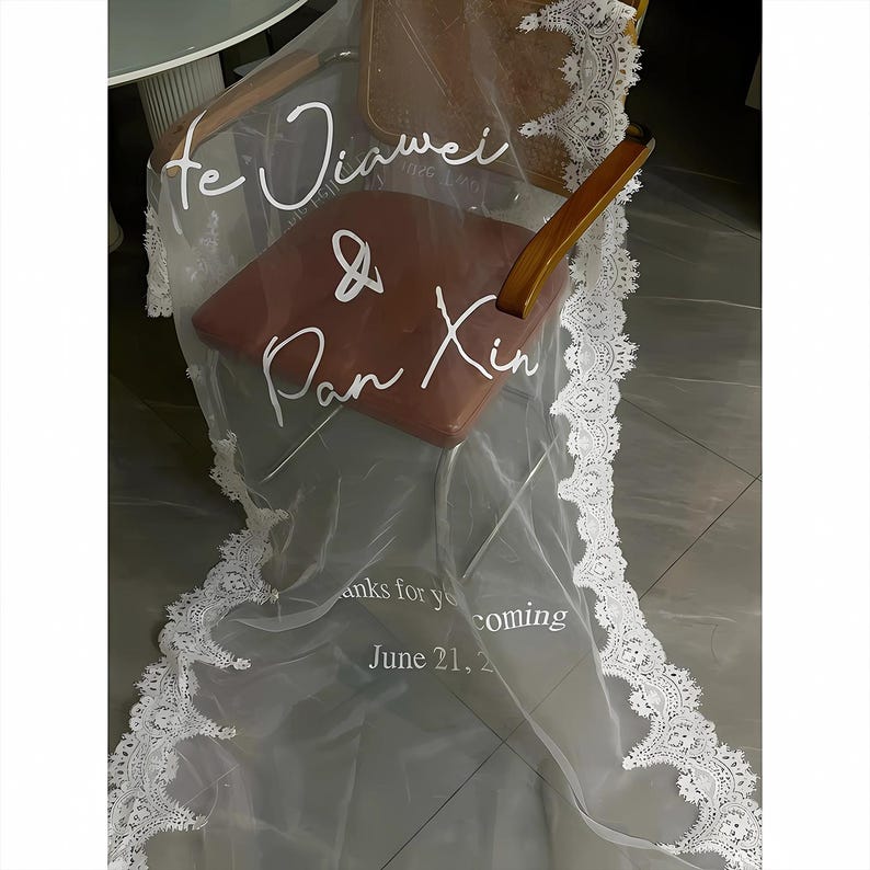 May include: A sheer, white wedding veil with a delicate lace trim. The veil is draped over a chair with a brown seat. Text on the veil reads "Te Jiawei & Pan Xin" and "June 21, 20_".