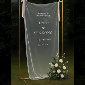 May include: A wedding welcome sign with the text "WELCOME TO THE WEDDING OF JENNY & YENKONG" on a sheer white fabric. The sign is held by a gold metal frame and decorated with white flowers and greenery. The date "May 10th 2025" is also displayed.