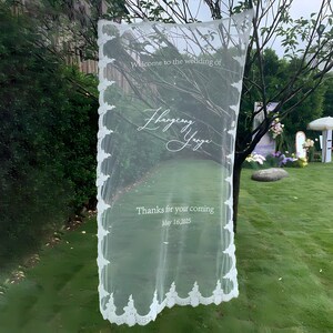 May include: A sheer white wedding sign with decorative lace edging. The sign reads "Welcome to the wedding of" followed by names and "Thanks for your coming May 16, 2025". The sign is hanging outdoors in front of a green background.