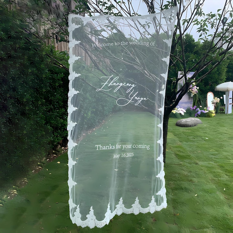 May include: A sheer white wedding sign with decorative lace edging. The sign reads "Welcome to the wedding of" followed by names and "Thanks for your coming May 16, 2025". The sign is hanging outdoors in front of a green background.