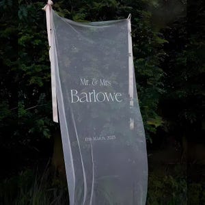 May include: A sheer white fabric wedding sign with the text "Mr. & Mrs Barlowe" and the date "12th March, 2025" in a delicate font. The sign is suspended with pink ribbons against a backdrop of green foliage.