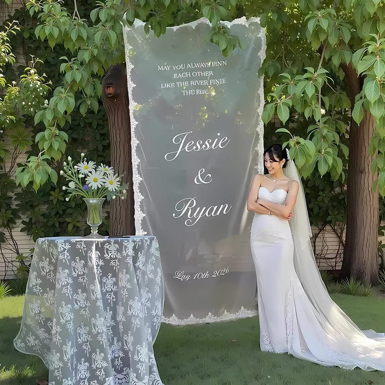May include: A wedding scene featuring a bride in a white strapless gown and veil. A sheer banner displays the names "Jessie & Ryan" and the date "Aug 10th 2026". A table with a floral arrangement and a lace tablecloth is also visible.