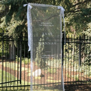 May include: A sheer wedding welcome sign with white lace trim and ribbons. The sign displays the text "WELCOME TO THE WEDDING OF ZANE & HANNAH" along with the date and location, August 29th, 2005, Salt Lake City, Utah.