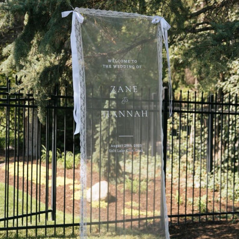 May include: A sheer wedding welcome sign with white lace trim and ribbons. The sign displays the text "WELCOME TO THE WEDDING OF ZANE & HANNAH" along with the date and location, August 29th, 2005, Salt Lake City, Utah.