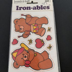 May include: A sheet of vintage "Iron-ables" scratch and sniff stickers. The stickers feature brown teddy bears playing a drum and holding a baseball bat, along with hearts and flowers. The packaging includes the text "Scratch & Sniff" and "1983 MORGAN INC."