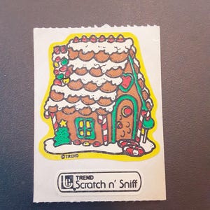 May include: A vintage "Scratch n' Sniff" sticker featuring a gingerbread house design. The house is brown with white frosting, candy cane accents, and a green Christmas tree. The sticker has a yellow border and the text "TREND".