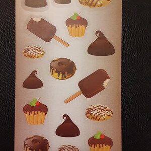 Peut inclure: Une planche d'autocollants avec des illustrations de desserts. Les autocollants représentent des bâtonnets glacés au chocolat, des cupcakes avec des cerises, des beignets au chocolat et des bonbons au chocolat. La palette de couleurs comprend des bruns, des beiges et du blanc.