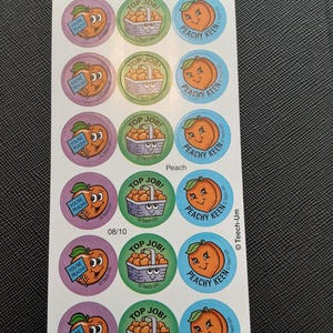 Puede incluir: Hoja de pegatinas redondas con diseños de melocotones. Las pegatinas presentan melocotones de dibujos animados con caras sonrientes, cestas de melocotones y frases como "Top Job!" y "Peachy Keen". Las pegatinas son de varios colores.