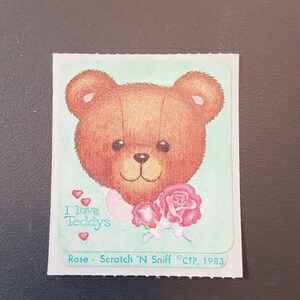 May include: A vintage sticker featuring a brown teddy bear with a smiling face, holding pink roses. The sticker has the text "I love Teddys" and "Rose - Scratch 'N Sniff ©CTP, 1983" on a light green background.