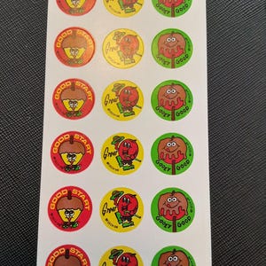 Puede incluir: Hoja de pegatinas vintage con personajes de dibujos animados. Las pegatinas presentan una bellota sonriente con "Good Start", una manzana roja con "Great" y una manzana marrón con "Goodey Good". Cada pegatina es redonda con un borde colorido.