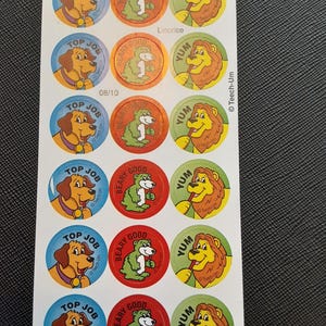 Puede incluir: Hoja de pegatinas coloridas con animales de dibujos animados. Los diseños incluyen un perro con "Top Job", un oso con "Beary Good" y un león con "Yum". Las pegatinas son redondas con bordes y texto de varios colores.