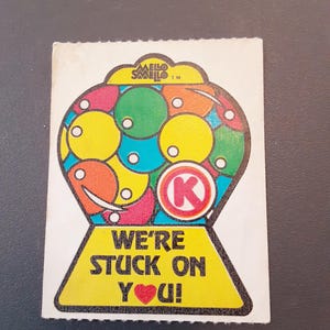 May include: A vintage sticker featuring a colorful gumball machine design. The machine is filled with various colored gumballs and has the text "WE'RE STUCK ON YOU!" with a red heart. The sticker also has the text "MELLO SMELLO" at the top.