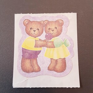 May include: A sticker featuring two brown teddy bears holding hands. One bear wears a yellow shirt and purple shorts, the other a yellow dress. They share a lollipop. The sticker has a light purple border and is on a white background.