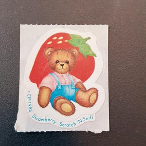 May include: A vintage sticker featuring a brown teddy bear wearing blue overalls, sitting in front of a red strawberry. The sticker has the text "Strawberry Scratch 'N Sniff" and "CTP 1983".