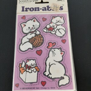 May include: Vintage "Iron-abs" scratch and sniff stickers featuring white cartoon cats with red hearts on a pink background. The sticker sheet includes cats playing with yarn, in a gift box, and looking at a butterfly. The packaging reads "Scratch & Sniff".