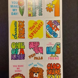 Könnte beinhalten: Ein Bogen mit Vintage-Aufklebern mit farbenfrohen Designs und Texten. Die Aufkleber zeigen Sätze wie "You Make My Day!", "You're Special!" und "Glad You're You!" mit Illustrationen von Blumen, Vögeln und Hunden.