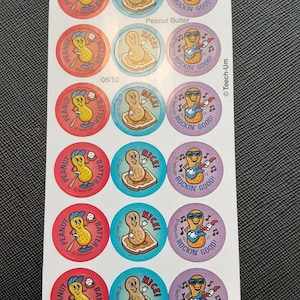 Puede incluir: Hoja de pegatinas redondas y coloridas con personajes de cacahuetes. Los diseños incluyen un cacahuete con un bate de béisbol, un cacahuete sobre una tostada y un cacahuete tocando la guitarra. El texto incluye "Peanut Batter", "Nice!" y "Rockin' Good!"