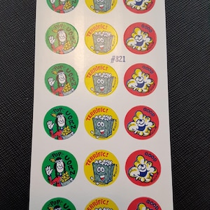 Puede incluir: Hoja de pegatinas de palomitas de maíz vintage. Las pegatinas redondas presentan personajes de dibujos animados de palomitas de maíz con los textos "Pop Corn", "Terrific!" y "Good". Las pegatinas son de varios colores, incluyendo verde, amarillo y rojo.