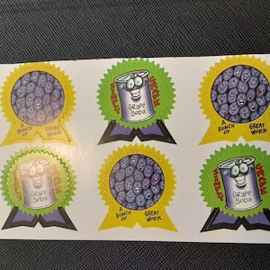 Puede incluir: Hoja de pegatinas con imágenes de dibujos animados. Algunas pegatinas muestran un círculo de caras moradas con el texto "A Bunch of Great Work". Otras presentan una lata sonriente de "Grape Soda".