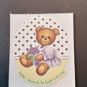 May include: A vintage sticker featuring a brown teddy bear holding a bouquet of purple flowers. The bear is wearing a lavender dress and is set against a dotted background. The text "Violet - Scratch 'N Sniff ©CTP, 1983" is printed on the sticker.