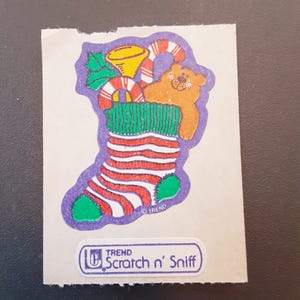 May include: A vintage Christmas-themed sticker featuring a stocking filled with a teddy bear, candy cane, and holly. The stocking is red and white striped with green accents. The sticker has the text "TREND Scratch n' Sniff".