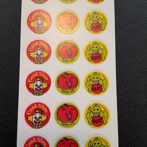 Puede incluir: Una hoja de pegatinas vintage con diseños de dibujos animados. Las pegatinas incluyen un payaso con el texto "Good Show", un tomate sonriente con el texto "Super Kid" y un caimán sonriente con el texto "Neat". Las pegatinas son redondas y de color rojo, amarillo y verde.
