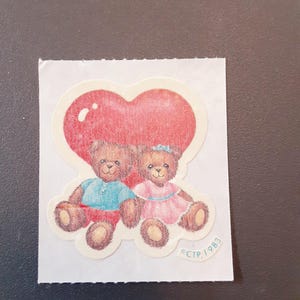 May include: A sticker featuring two teddy bears under a red heart. One bear wears a blue shirt and red shorts, the other a pink dress. The sticker has a white border and the text "CTP, 1983".