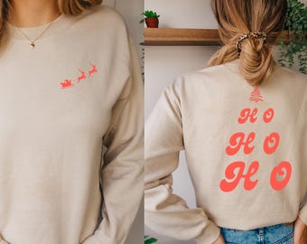 May include: Beige sweatshirt with a red Santa and reindeer graphic on the front. The back of the sweatshirt features a red Christmas tree design with the words "Ho Ho Ho". The sweatshirt is a long-sleeved crew neck.