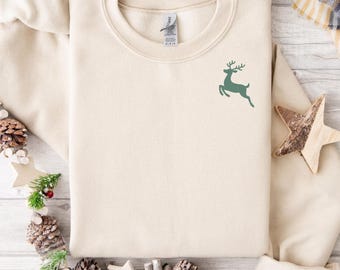 May include: A cream-coloured sweatshirt featuring a green deer graphic on the upper left chest. The sweatshirt is neatly folded on a wooden surface, surrounded by pine cones, stars, and greenery. A plaid blanket is visible in the background.