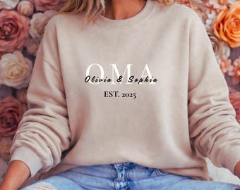 May include: A beige sweatshirt with the word "OMA" in white, and the names "Olivia & Sophia" in a script font. Below the names is the text "EST. 2025" in a simple font. The sweatshirt is a neutral color.