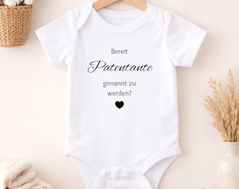 Puede incluir: Body de bebé blanco con el texto en alemán "Bereit Patentante genannt zu werden?" en escritura negra, con un corazón negro. La prenda está colgada en una percha de madera.
