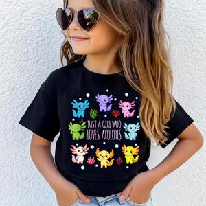 Könnte beinhalten: Schwarzes T-Shirt mit bunten Axolotl-Grafiken und dem Text "JUST A GIRL WHO LOVES AXOLOTLS." Das Design zeigt Axolotl in Blau, Lila, Grün, Rosa, Orange und Gelb. Das T-Shirt wird von einer jungen Person mit herzförmiger Sonnenbrille getragen.