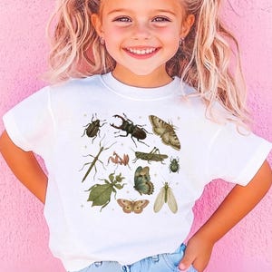Puede incluir: Una camiseta blanca de manga corta con un estampado gráfico detallado de varios insectos, incluyendo escarabajos, una polilla, una mariposa, una mantis religiosa y un insecto palo, dispuestos en grupo con pequeñas estrellas.