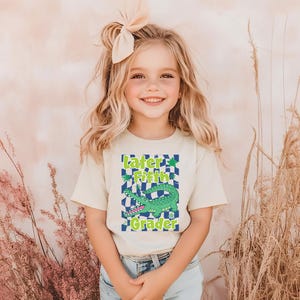 May include: A young girl wearing a cream-colored t-shirt with a graphic that says "Later Fifth Grader" and features a green alligator on a checkered background. She is smiling and wearing a bow in her hair. The background is a soft, neutral color.