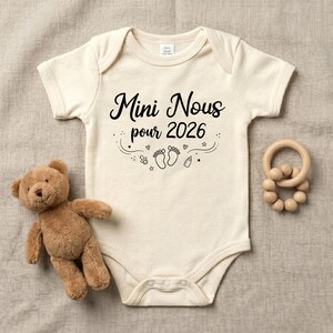 May include: A cream-colored short-sleeve baby bodysuit featuring black text that reads Mini Nous pour 2026, accompanied by graphic illustrations of baby footprints, a pacifier, a baby bottle, small hearts, and stars, with a brown teddy bear.