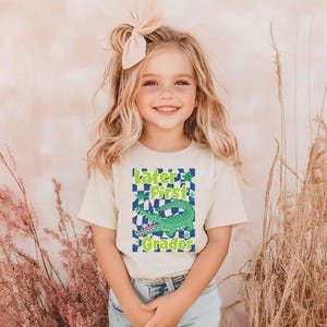 May include: A light beige t-shirt with the text "Later First Grader" and a green alligator graphic. The shirt is worn by a young girl with blonde hair and a peach-colored bow.