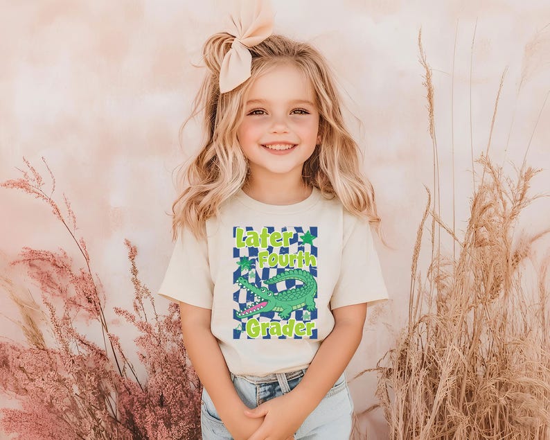 May include: A young girl wearing a cream-colored t-shirt with a graphic that says "Later Fourth Grader." The graphic features a green alligator on a blue and white checkered background. She is also wearing blue jeans and a cream-colored bow.
