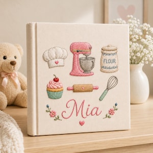 Könnte beinhalten: Ein quadratisches, cremefarbenes, stoffbezogenes Buch mit gestickten Backmotiven, darunter eine Kochmütze, eine rosa Küchenmaschine, ein Mehlbehälter, ein Cupcake, ein Nudelholz und ein Schneebesen. Der Name Mia ist in rosa Schrift gestickt.