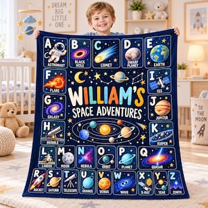 May include: A dark blue fleece blanket featuring an alphabet-themed space design. The center displays the text "WILLIAM'S SPACE ADVENTURES" above a solar system graphic. Each letter from A to Z is paired with a space-related illustration and label.