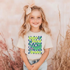 May include: A light beige t-shirt with the text "Later Third Grader" in green and blue, featuring a cartoon alligator. The shirt is worn by a young girl with blonde hair and a bow.