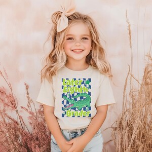 May include: A young girl wearing a cream-colored t-shirt with a graphic design. The design features the text "Later Eighth Grader" above a green alligator on a checkered background. She is also wearing blue jeans and a bow in her hair.