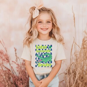 May include: A light beige t-shirt with the text "Later Seventh Grader" and a green alligator graphic. The shirt is worn by a young girl with blonde hair and a bow. The background is a light pink.