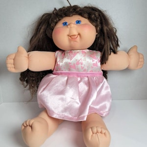 May include: A vintage Cabbage Patch Kid doll with brown hair and blue eyes. The doll is wearing a pink satin dress with a white bodice decorated with pink hearts. The doll is giving a thumbs up with both hands.