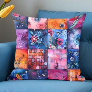 May include: Square cushion with a patchwork design. The cushion features a variety of colourful squares with floral and abstract patterns in shades of blue, orange, pink, and purple. The cushion is placed on a blue sofa.