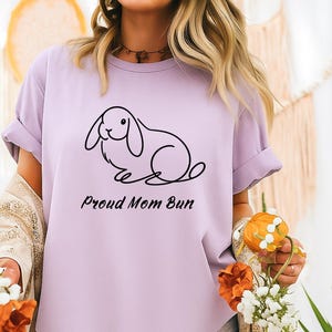 May include: A lilac-colored t-shirt featuring a minimalist black line drawing of a rabbit. The text "Proud Mom Bun" is printed below the rabbit illustration. The shirt has short sleeves and a relaxed fit.
