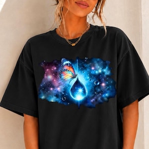 May include: A black short-sleeve t-shirt featuring a graphic print of a colorful butterfly perched on a glowing blue water droplet. The background of the print depicts a vibrant nebula with swirling shades of purple, blue, and scattered white stars.