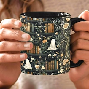 May include: A black ceramic mug featuring a dark, intricate pattern of bookshelves filled with various books, interspersed with white ghost figures and clusters of small, light-colored flowers. The interior and handle of the mug are solid black.